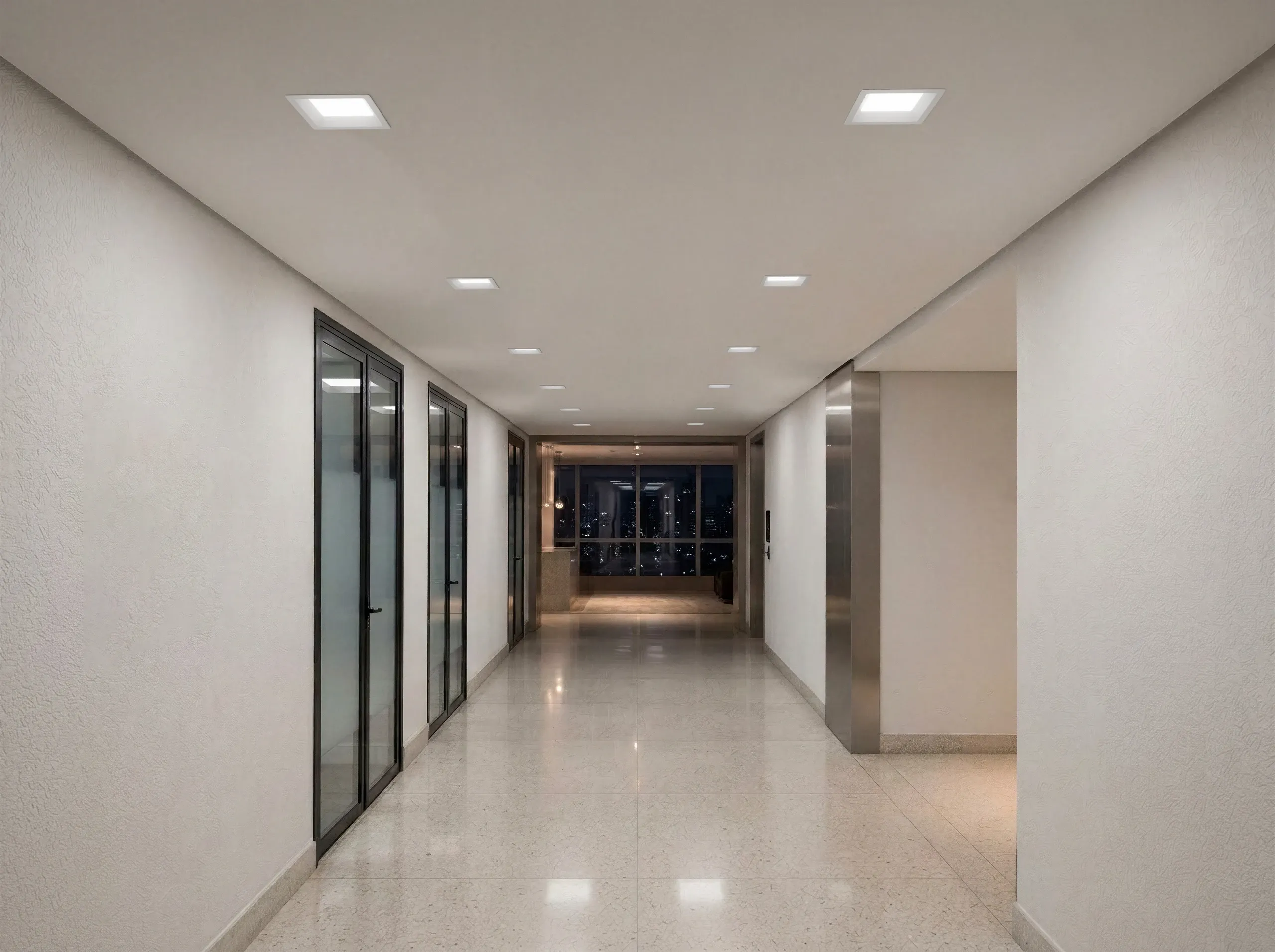 Corredor de prédio à noite iluminado por downlights Hide quadrados embutidos no forro branco, com piso de granilite polido refletindo a luz e vista da cidade ao fundo.